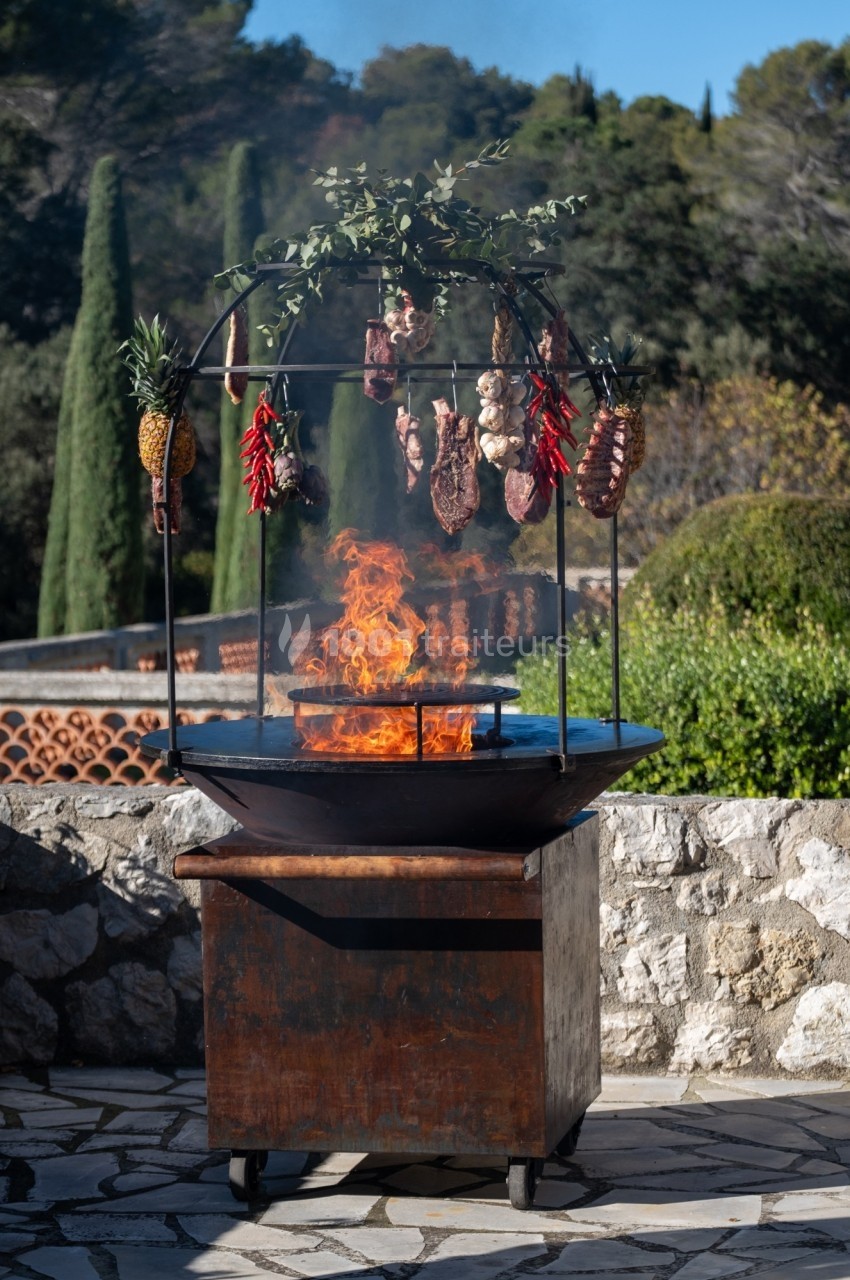 Grill extérieur avec feu de bois, viandes, ananas et piments suspendus, entouré de végétation méditerranéenne.