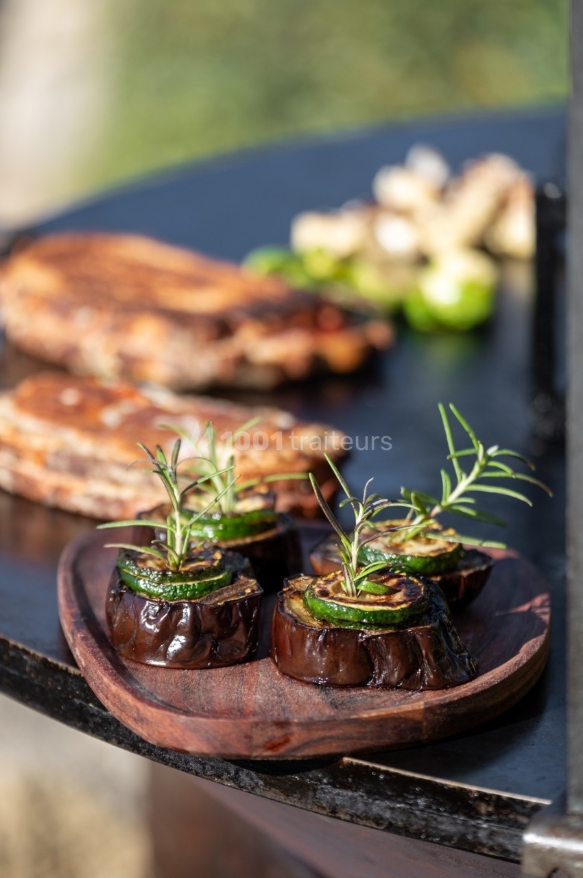 Tranches d'aubergines grillées garnies de romarin sur une assiette en bois, avec des aliments cuits en arrière-plan.