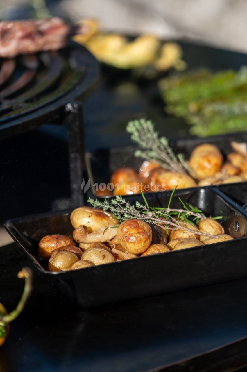 Pommes de terre rôties dans un plat en fonte, garnies de branches de thym, avec un barbecue en arrière-plan.