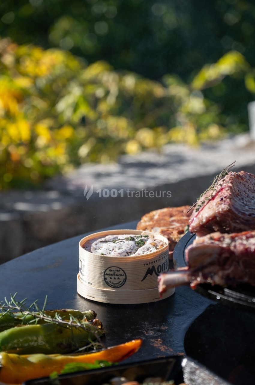 Fromage Mont d'Or fondu dans sa boîte en bois, accompagné de légumes grillés et de viande, en extérieur.