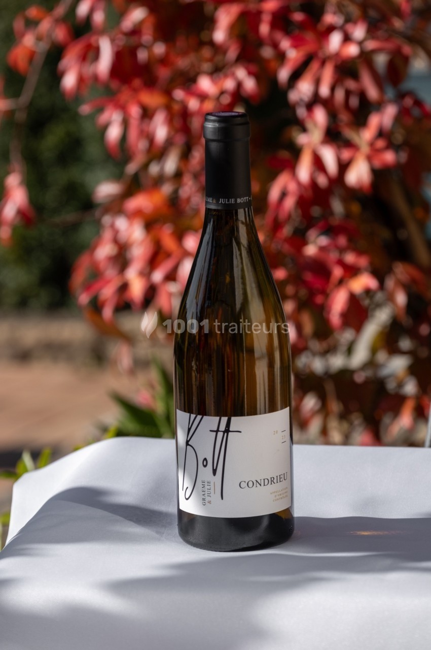 Bouteille de vin blanc posée sur une table blanche, avec un fond flou de feuillage rouge et vert.