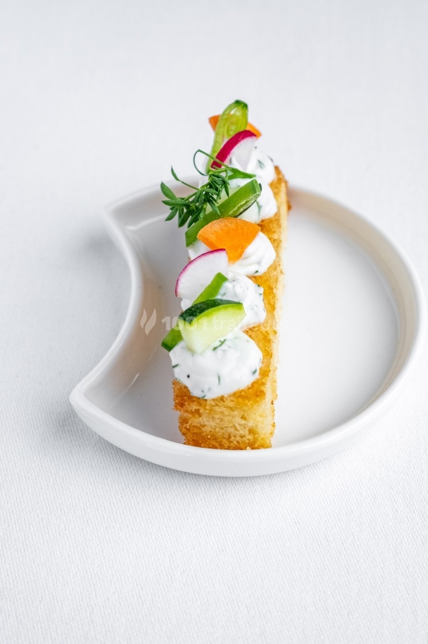 Tranche de pain doré garnie de crème, herbes fraîches et légumes colorés, présentée sur une assiette blanche.