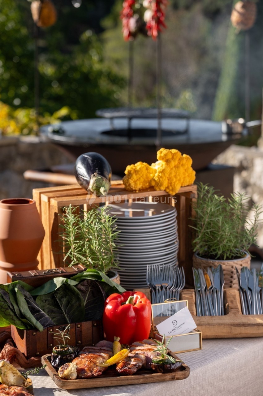 Table dressée en extérieur avec des plats grillés, légumes frais, herbes aromatiques et un barbecue en arrière-plan.