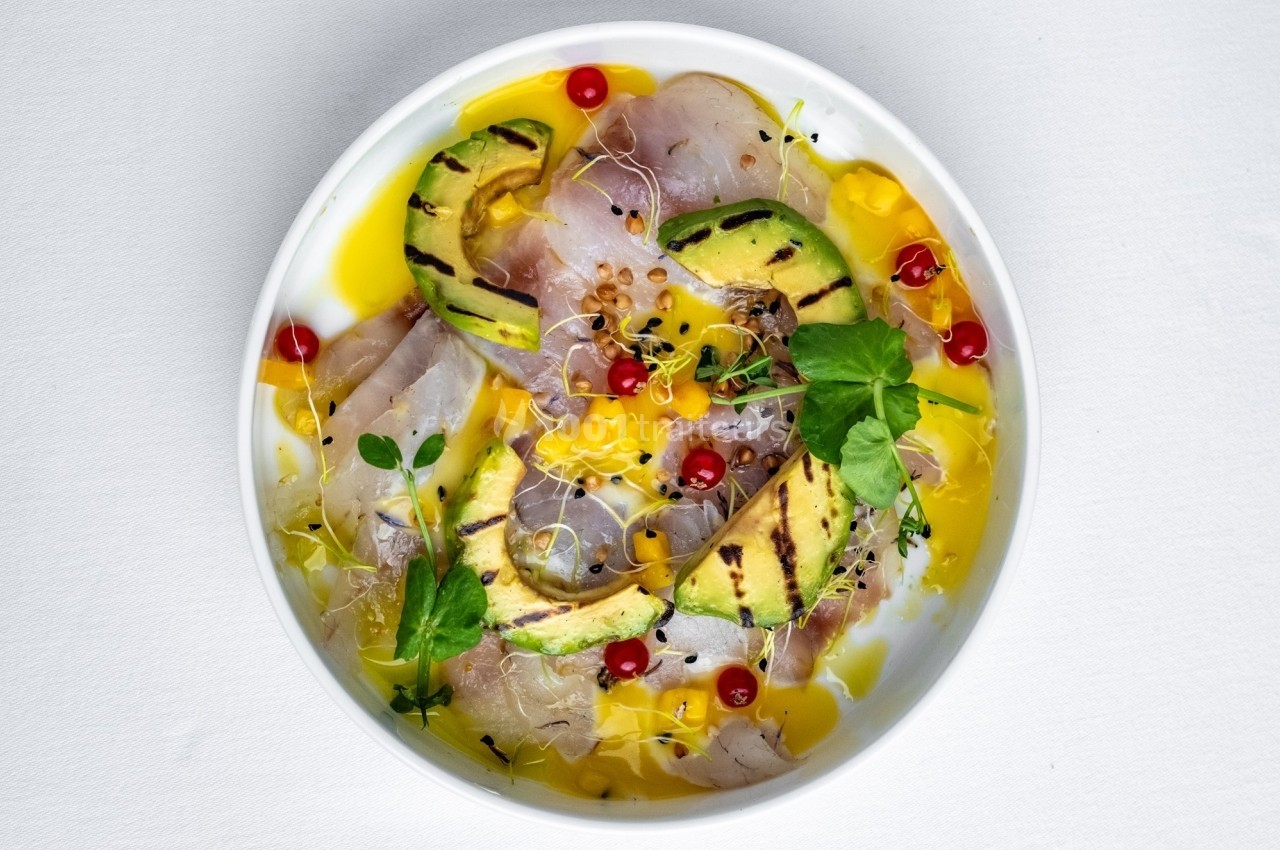 Assiette de carpaccio de poisson avec tranches d'avocat grillé, groseilles rouges, pousses et sauce jaune.