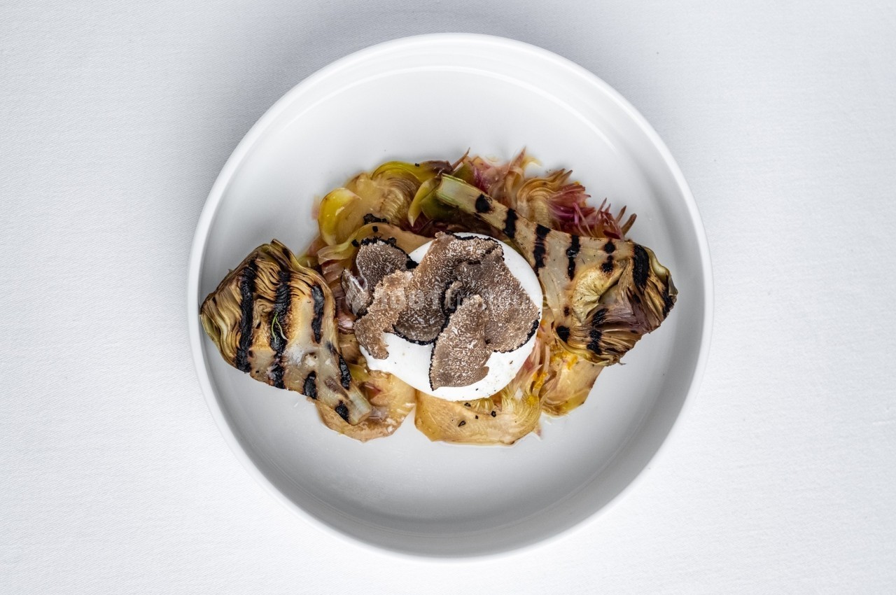 Assiette contenant de la burrata, des tranches de truffe et des artichauts grillés, présentée sur une nappe blanche.