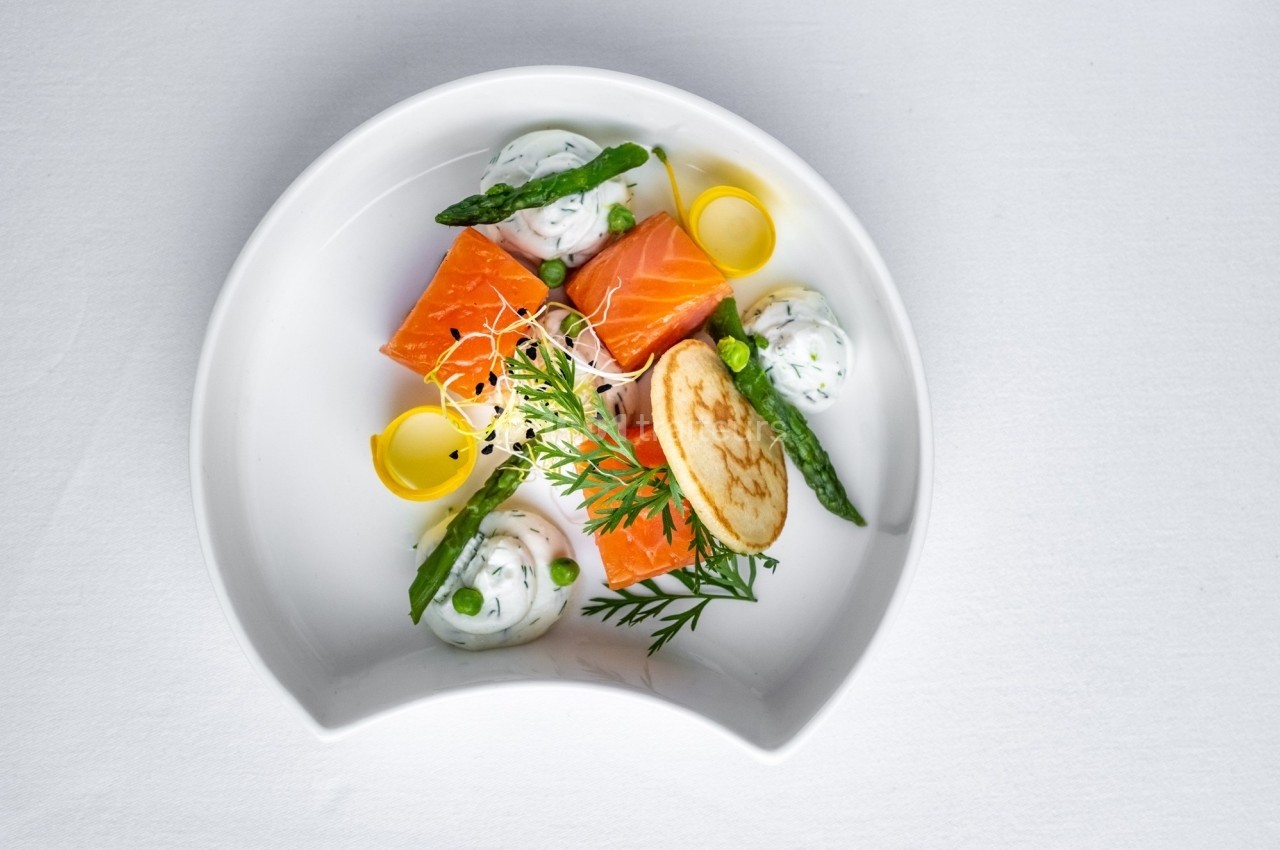 Assiette gastronomique avec saumon, asperges, crème aux herbes, blinis et décorations végétales sur fond blanc.