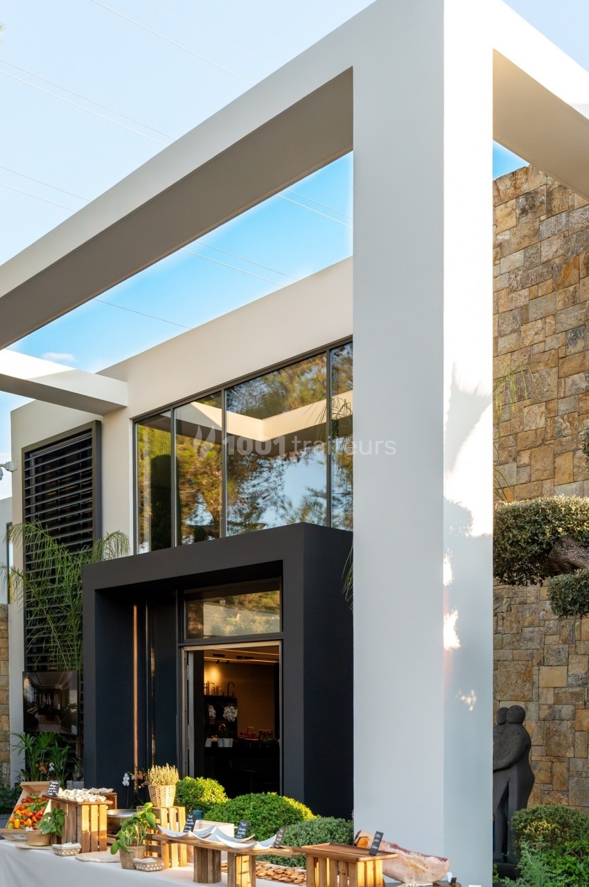 Façade moderne d'une maison avec grandes baies vitrées, structure géométrique et table de buffet en extérieur.
