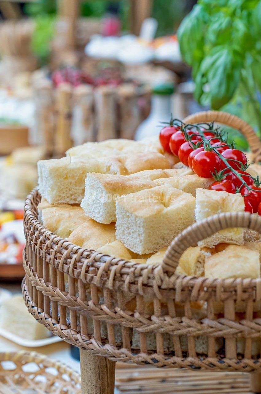 Panier en osier contenant des morceaux de focaccia et des tomates cerises sur une table garnie de plats variés.