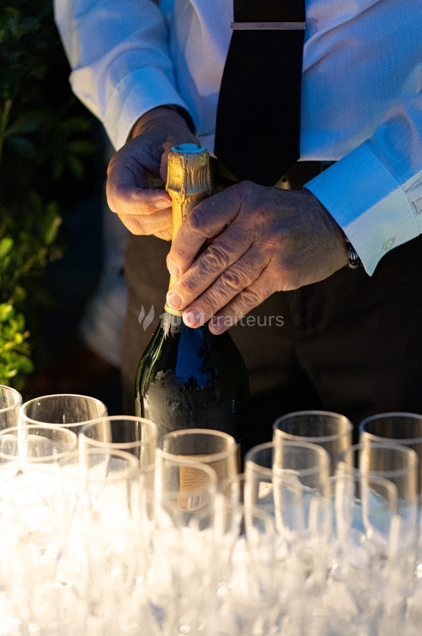 Un serveur ouvre une bouteille de champagne devant une rangée de flûtes vides sur une table éclairée.