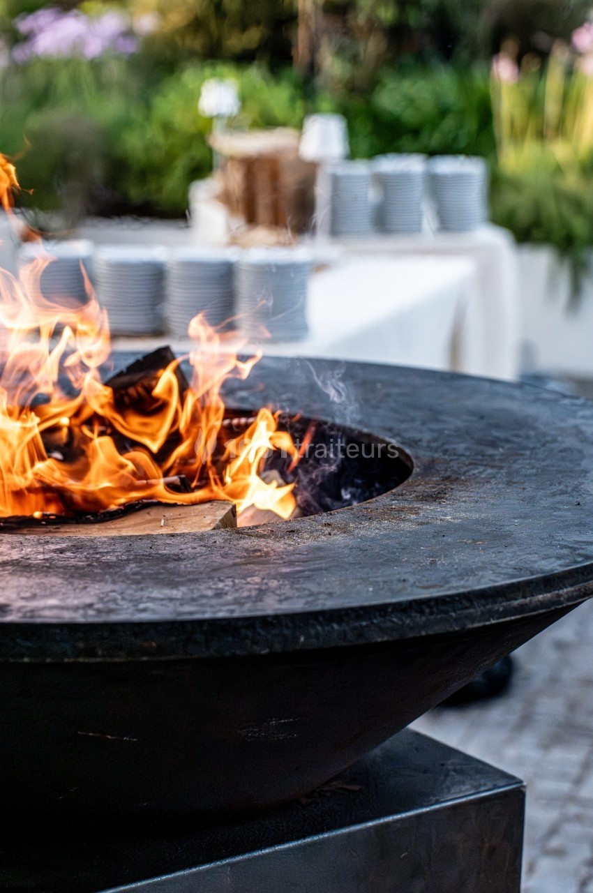 Brasero en métal noir avec des flammes vives, en arrière-plan une table dressée avec des assiettes empilées.