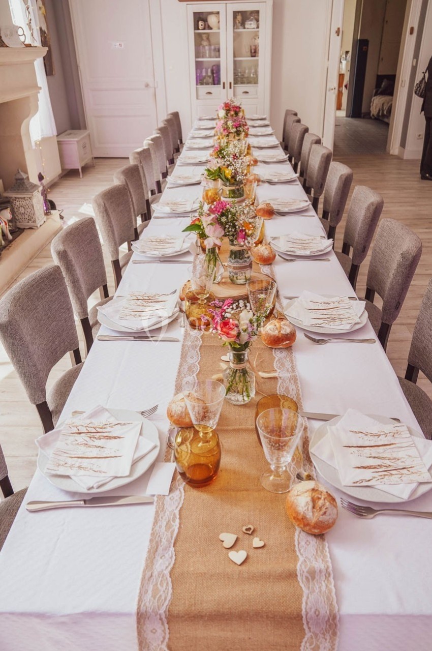 Table dressée pour un repas, décorée avec un chemin de table en toile de jute, fleurs, vaisselle et petits pains.