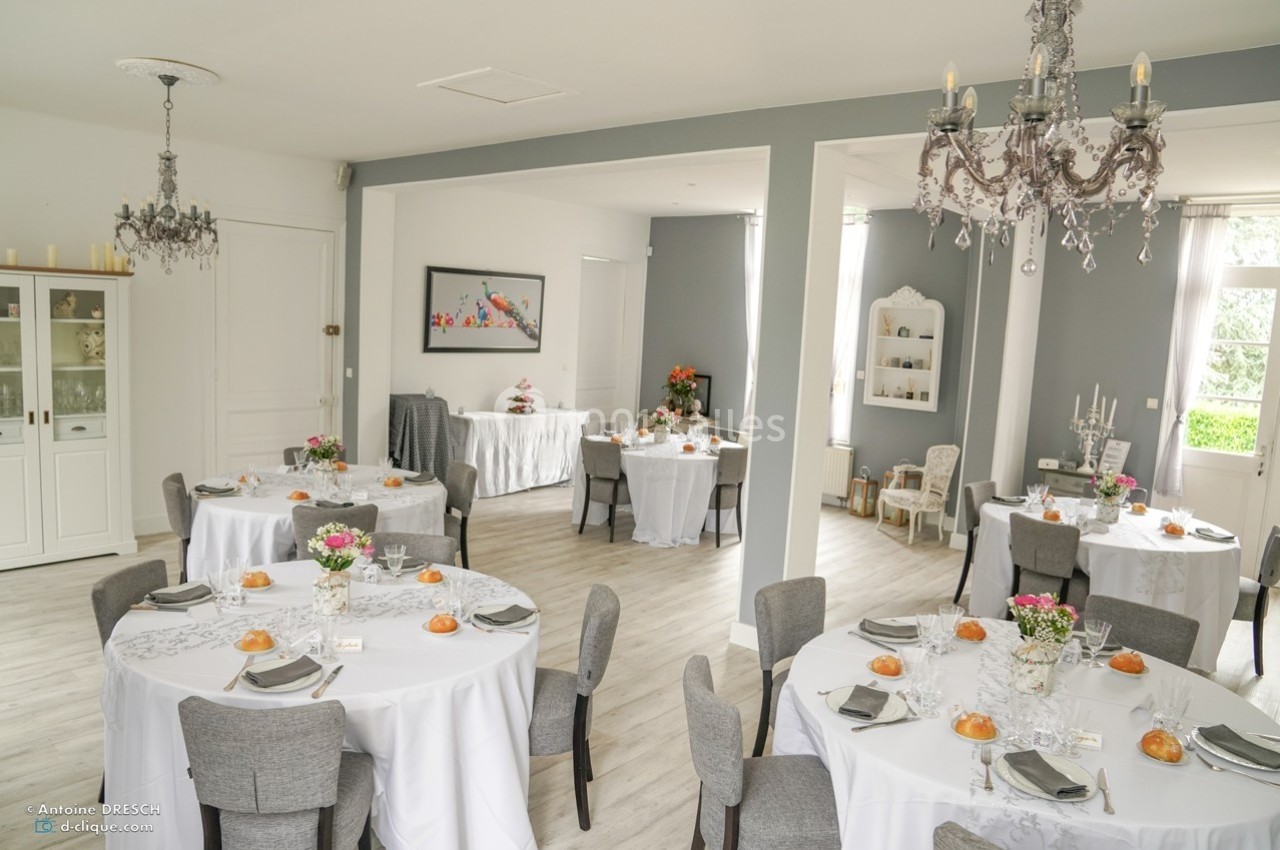 Salle de réception lumineuse avec tables rondes dressées, décorée de fleurs et éclairée par des lustres.