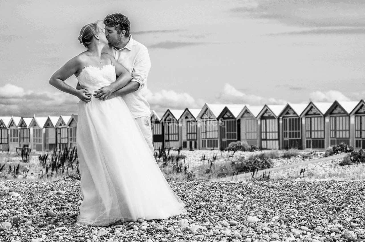 Un couple en tenue de mariage s'embrasse sur une plage de galets devant des cabines alignées.