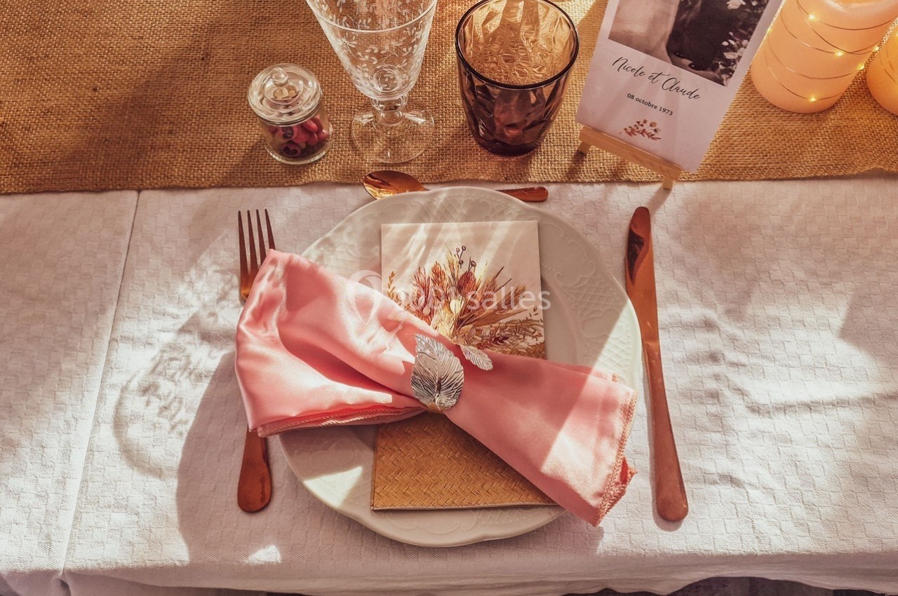 Table dressée avec une assiette blanche, une serviette rose ornée d'une feuille argentée, et des couverts cuivrés.