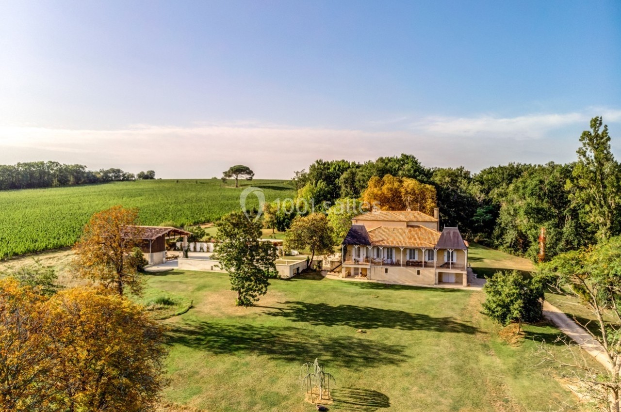 Vue aérienne d'une grande maison entourée de jardins, d'arbres et de champs sous un ciel dégagé.