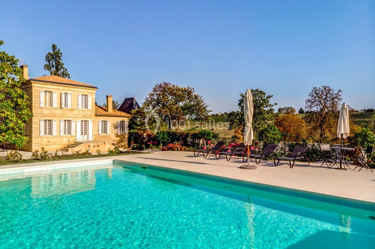 Piscine extérieure bordée de chaises longues et parasols, avec une maison en pierre et un jardin en arrière-plan.