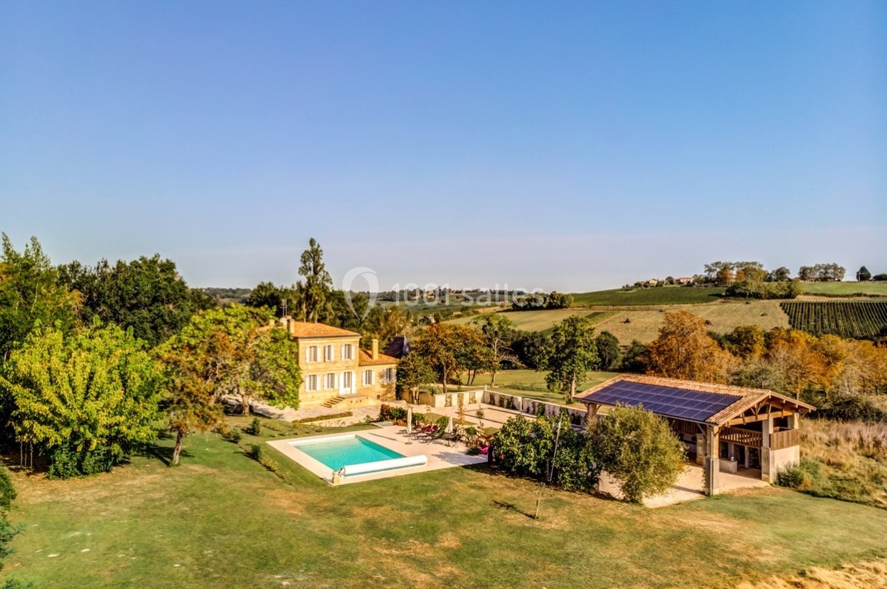 Vue aérienne d'une maison en pierre avec piscine, entourée de jardins et de champs sous un ciel dégagé.