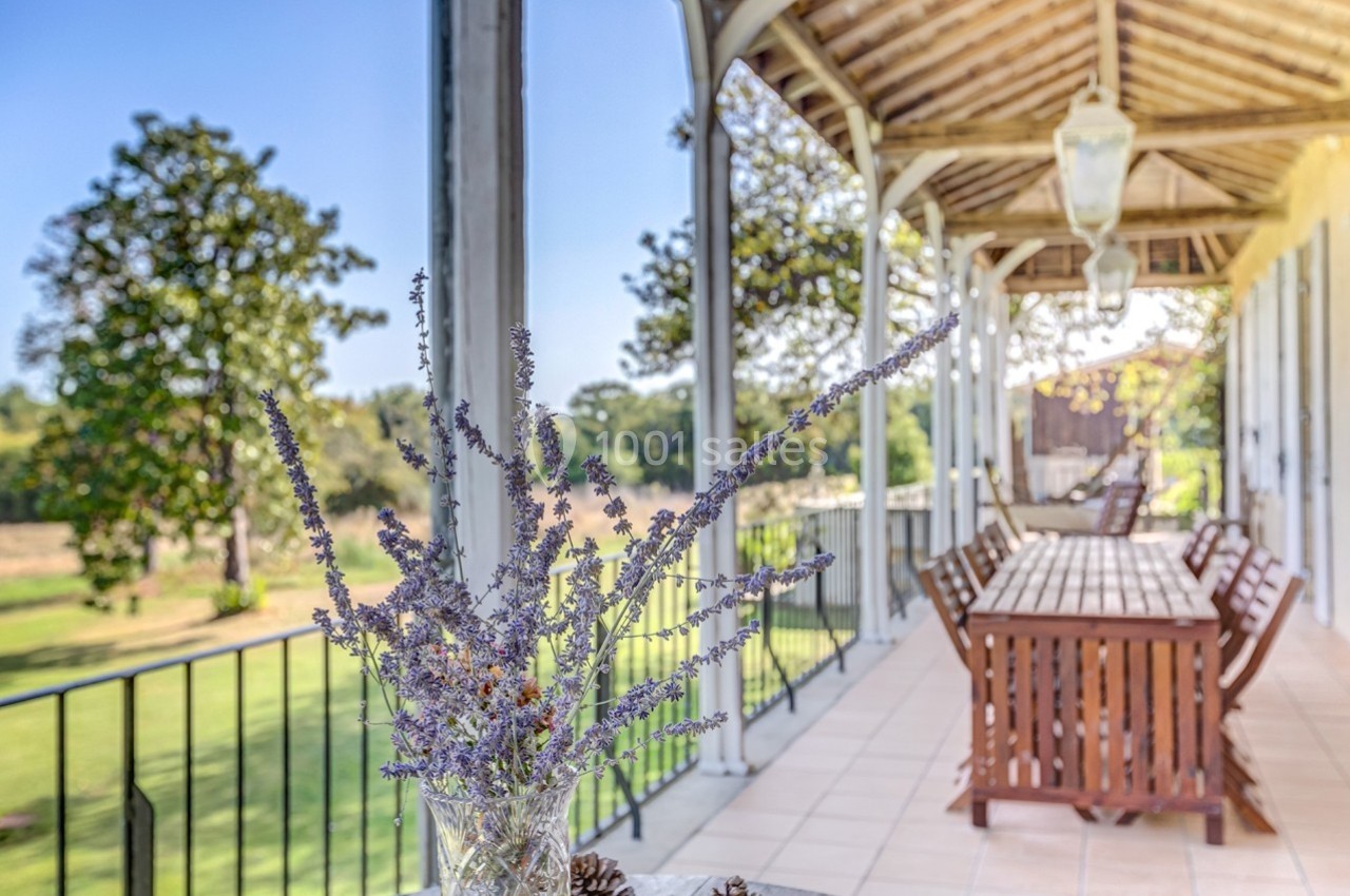 Terrasse couverte avec table en bois, vue sur un jardin verdoyant et un vase de fleurs séchées au premier plan.