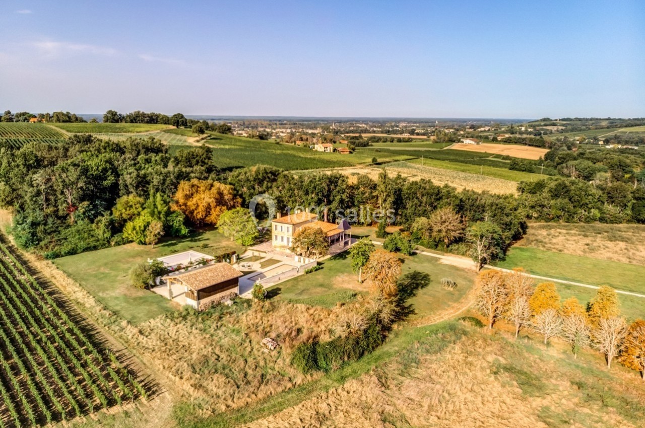 Vue aérienne d'une maison entourée de vignes, de champs et d'arbres dans un paysage rural ensoleillé.