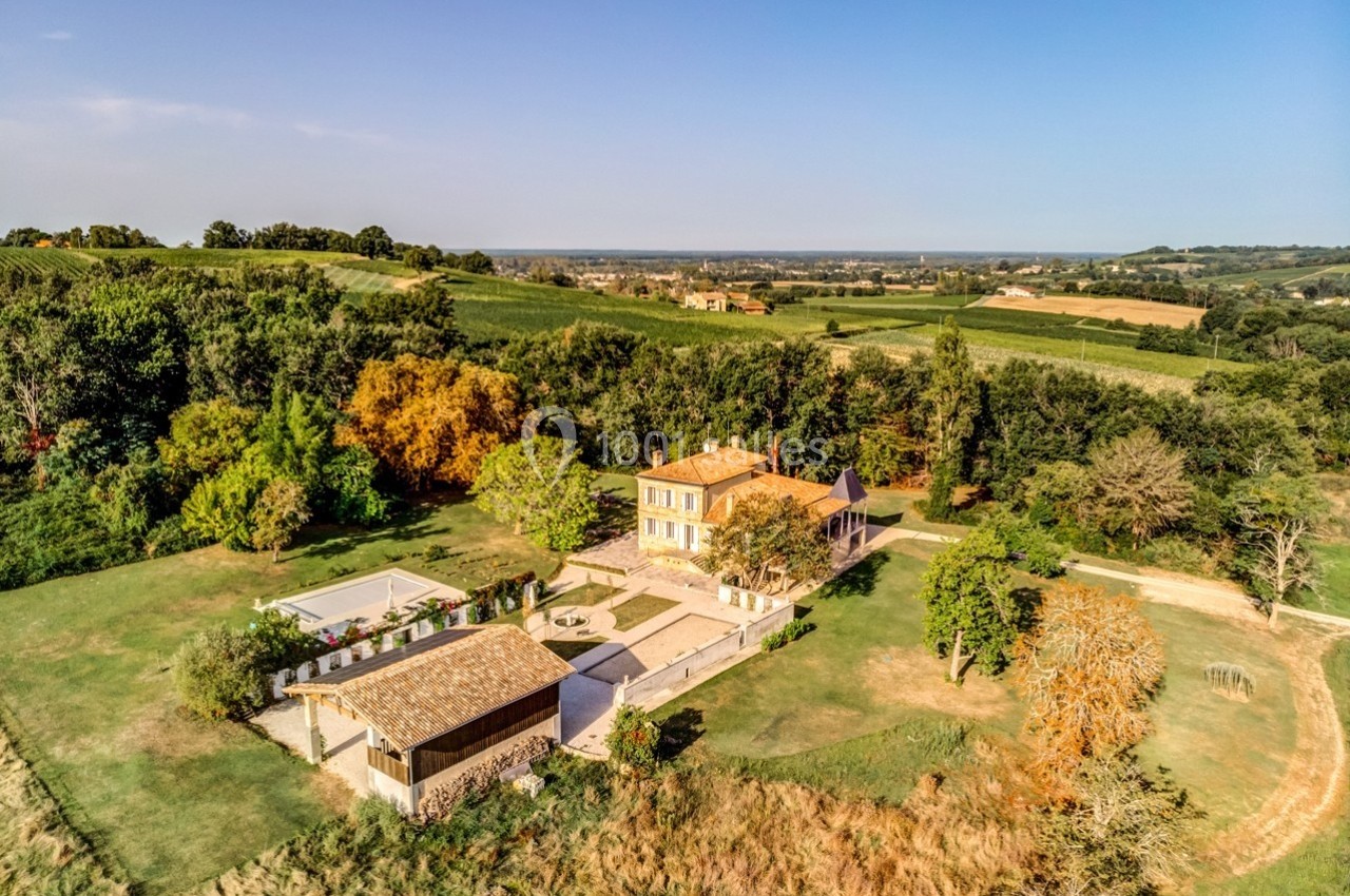 Vue aérienne d'une maison entourée de jardins, d'arbres et de champs dans un paysage rural ensoleillé.