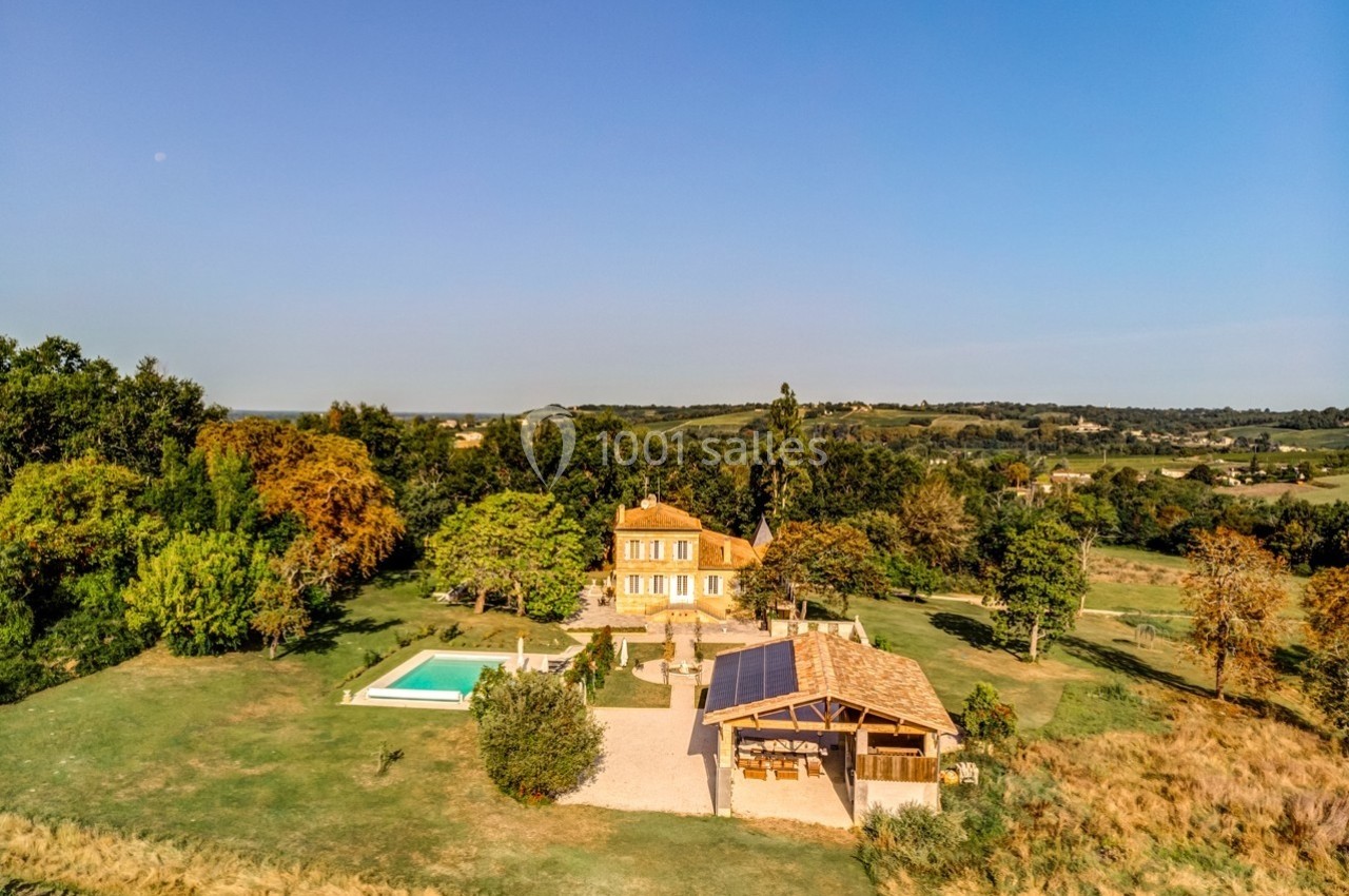 Vue aérienne d'une maison avec piscine, terrasse et jardin arboré, entourée de champs et collines sous un ciel dégagé.