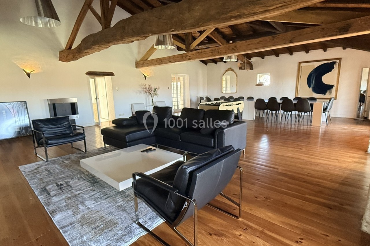 Salon spacieux avec poutres apparentes, canapé en cuir noir, table basse blanche et salle à manger en arrière-plan.