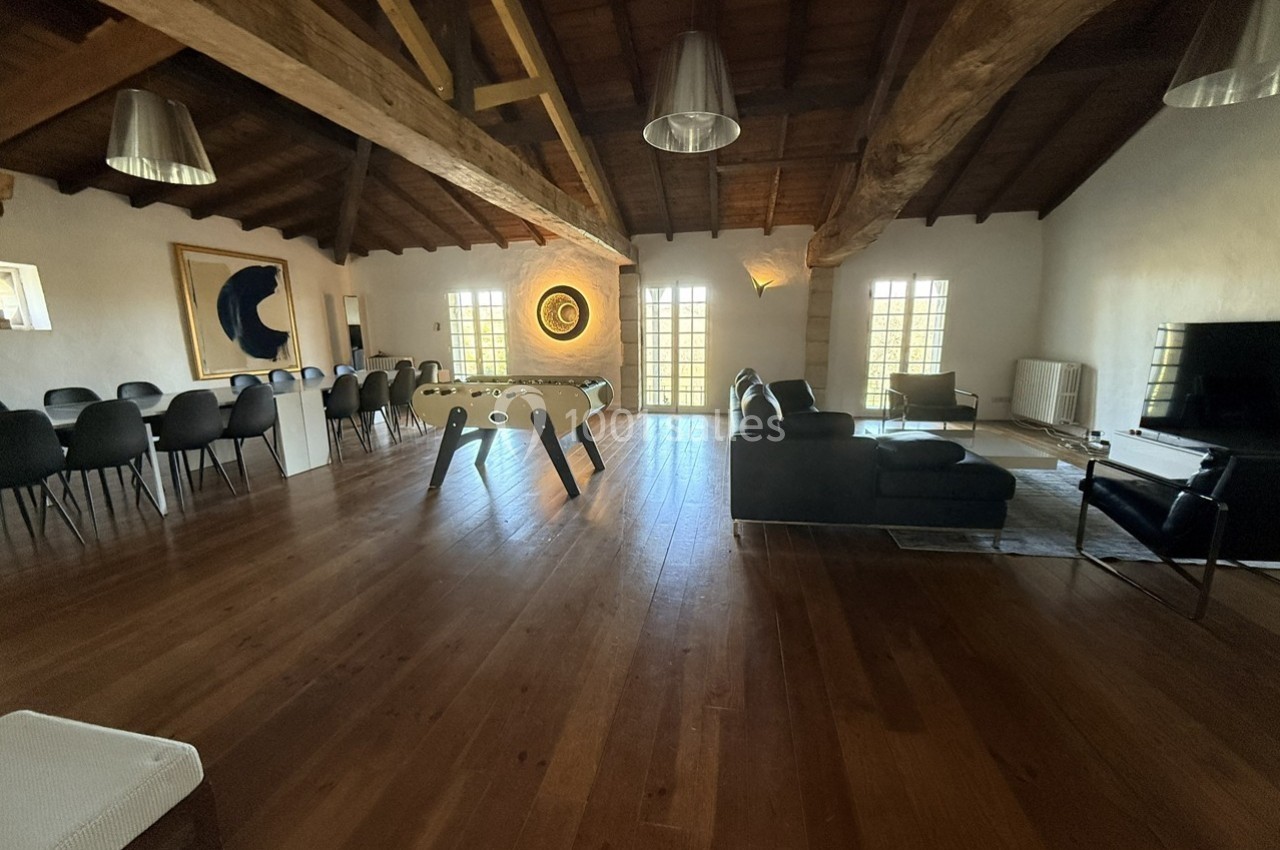 Salle spacieuse avec parquet, table de baby-foot, coin salon, grande table à manger et poutres apparentes au plafond.