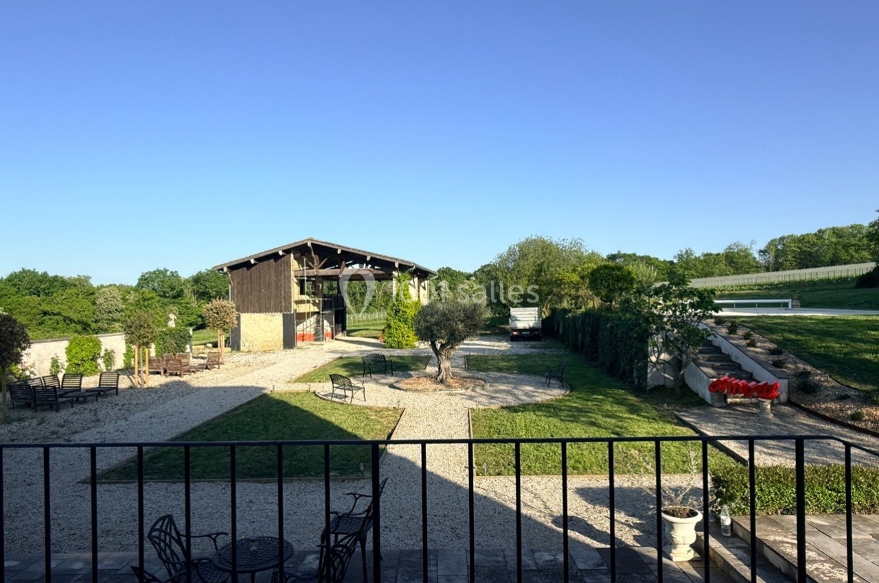 Vue d'une cour extérieure avec une grange, des espaces verts, des arbres et des meubles de jardin sous un ciel dégagé.