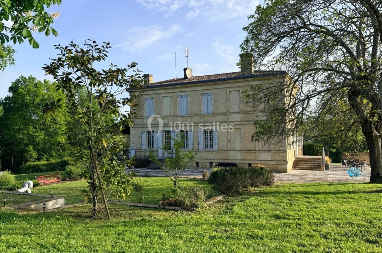 Grande maison en pierre entourée d'un jardin verdoyant avec arbres, pelouse et mobilier d'extérieur.