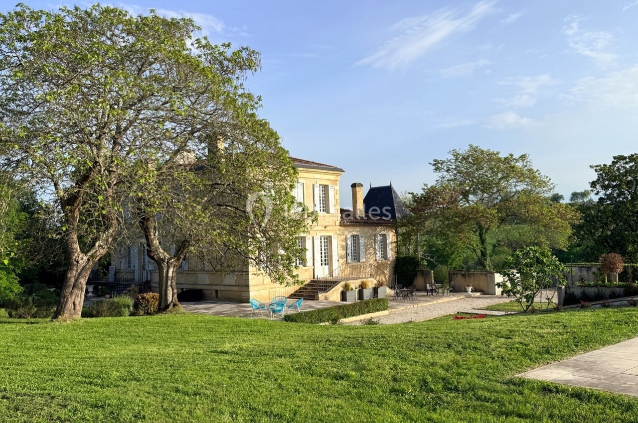 Manoir en pierre entouré de pelouse, arbres et terrasse, sous un ciel dégagé en journée.