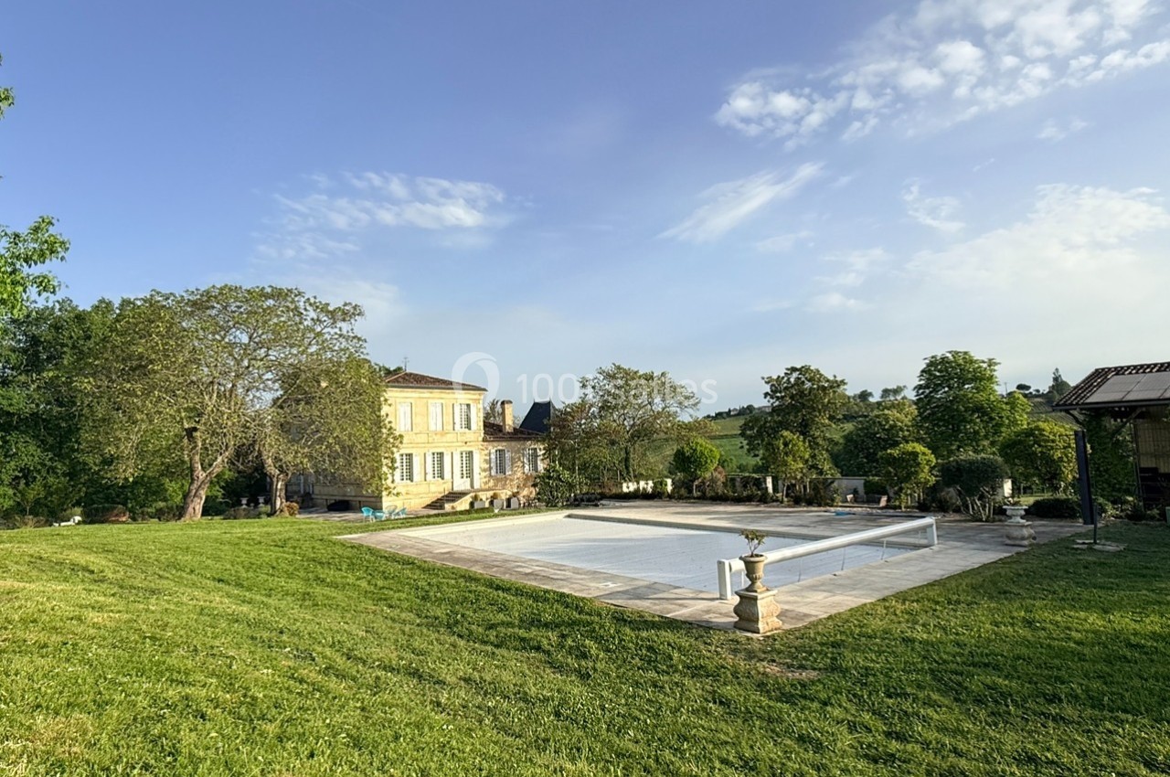 Grande maison en pierre entourée de verdure avec une piscine couverte au premier plan sous un ciel dégagé.