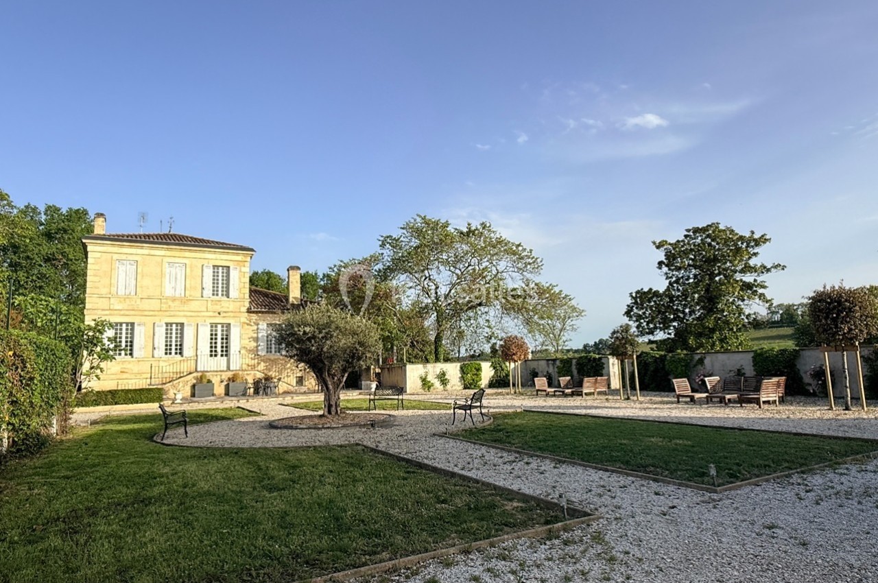 Jardin avec allées en gravier, pelouse, arbres et mobilier d'extérieur, devant une grande maison en pierre.