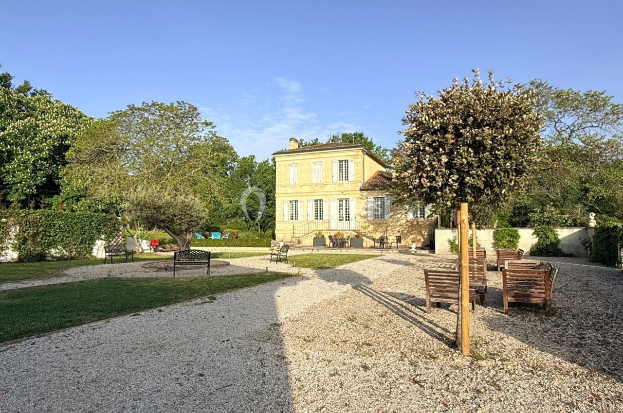 Maison en pierre jaune entourée d'un jardin avec arbres, bancs et allées en gravier sous un ciel dégagé.