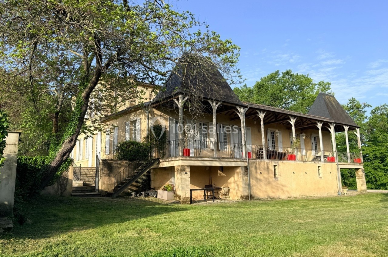 Grande maison ancienne avec balcon en bois, toit en pente et jardin arboré sous un ciel dégagé.