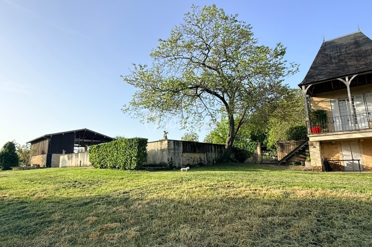 Grand jardin avec pelouse, arbre central, bâtiment en pierre à droite et hangar en arrière-plan sous un ciel dégagé.