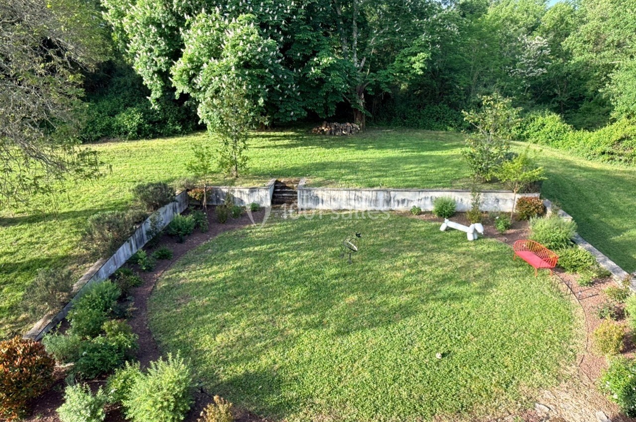 Jardin circulaire avec pelouse, bordé de plantes et arbustes, entouré d'un muret, avec des arbres en arrière-plan.