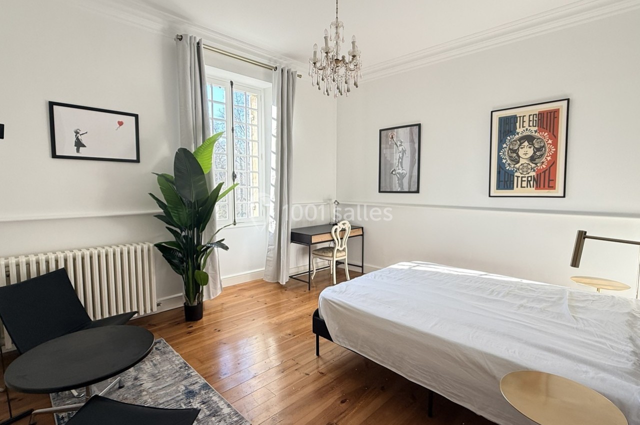 Chambre lumineuse avec lit, bureau, plantes, parquet en bois, tableaux encadrés et lustre suspendu.
