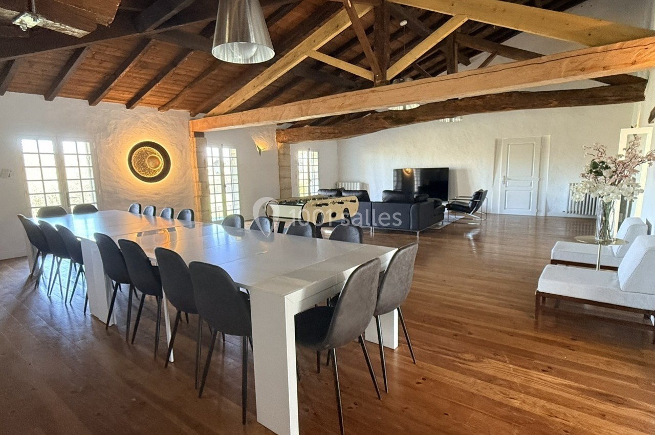 Grande salle lumineuse avec table à manger, chaises noires, canapé, baby-foot et poutres apparentes au plafond.