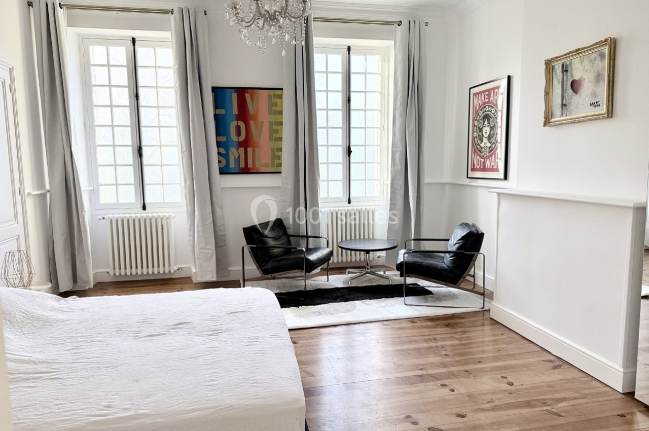 Chambre lumineuse avec parquet, lit blanc, fauteuils noirs, table basse, et décorations murales colorées.