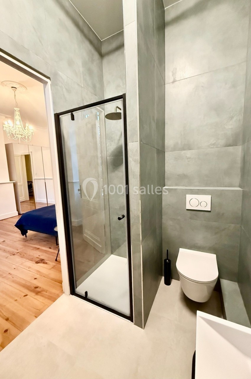 Salle de bain moderne avec douche vitrée, toilettes suspendues et murs en carrelage gris, vue sur une chambre adjacente.