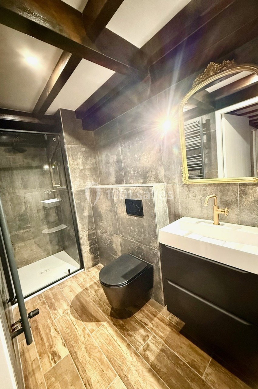 Salle de bain moderne avec douche à l'italienne, lavabo avec miroir doré, toilettes noires et sol en bois.