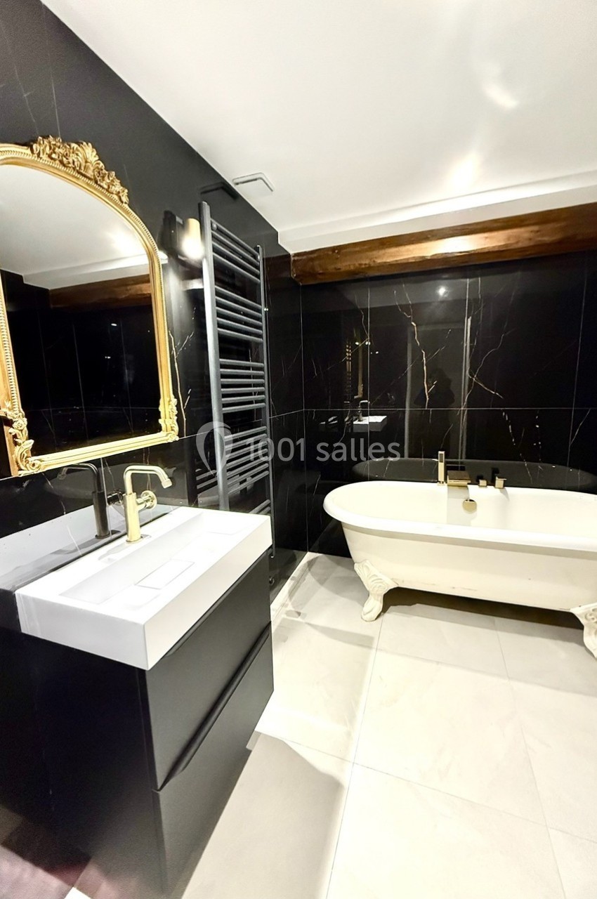 Salle de bain moderne avec murs noirs, baignoire sur pieds, miroir doré et lavabo blanc sur meuble noir.