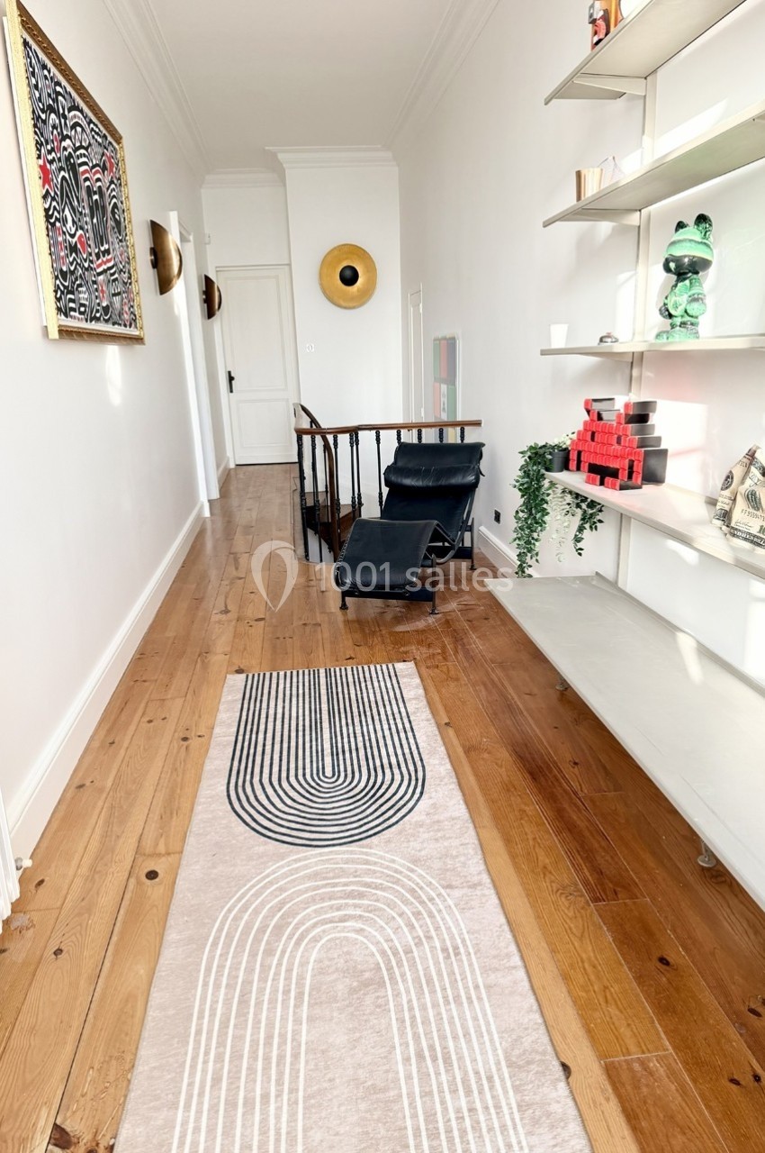 Couloir lumineux avec parquet, tapis à motifs géométriques, étagères décorées et fauteuil noir près d'un escalier.