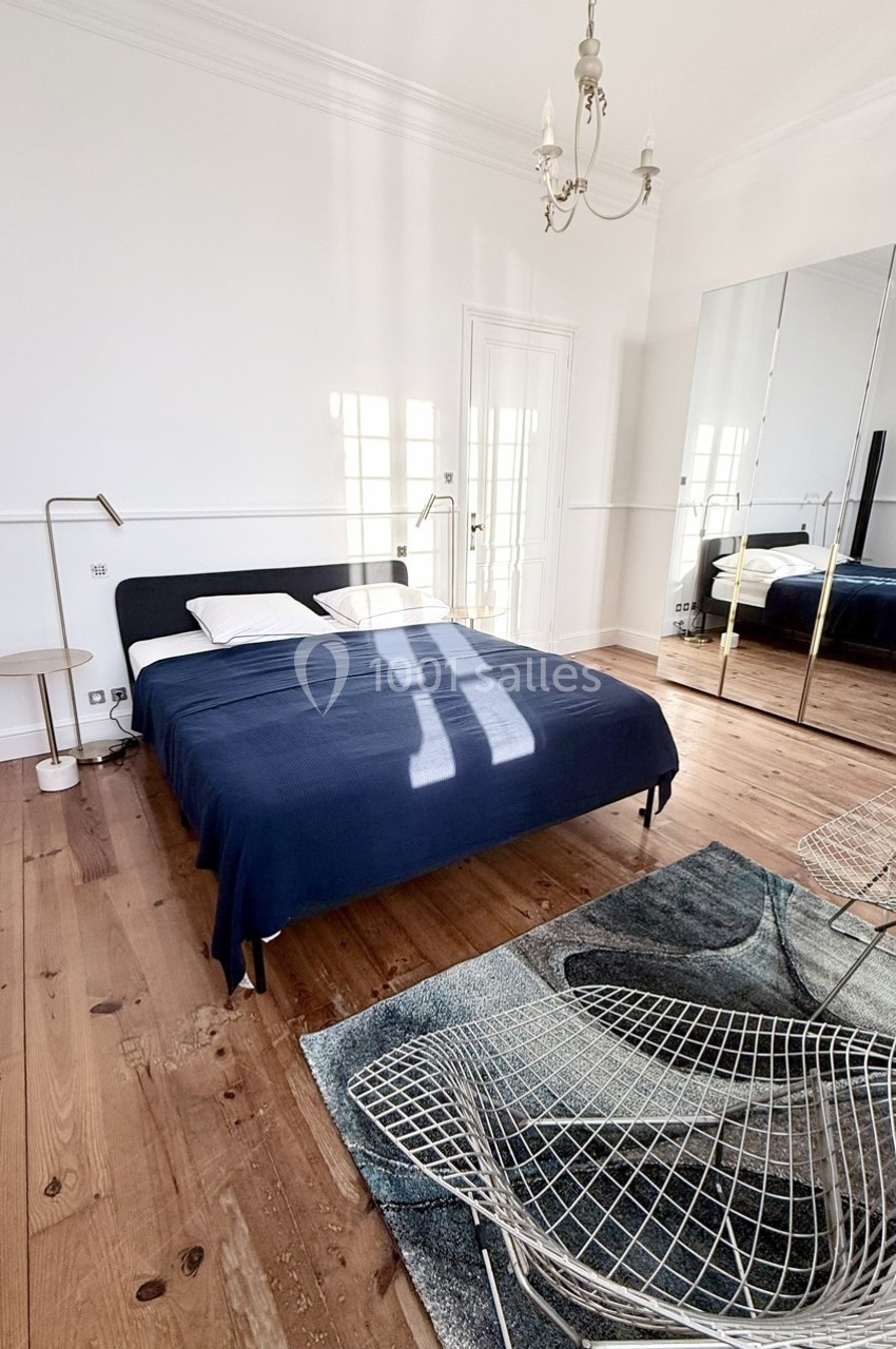 Chambre lumineuse avec lit double, couverture bleue, parquet en bois, miroir, et chaise en métal sur un tapis.