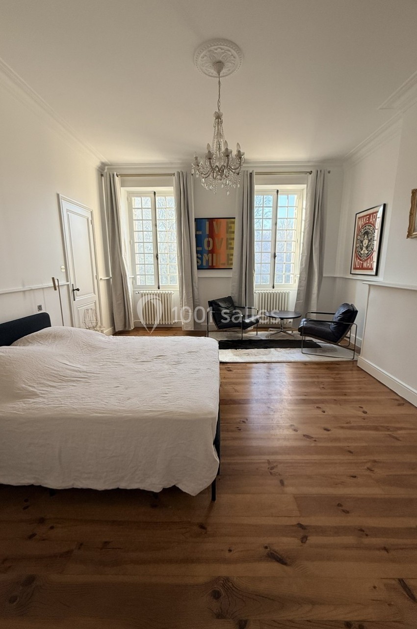 Chambre lumineuse avec lit double, parquet en bois, deux fauteuils noirs, tapis clair et grandes fenêtres.