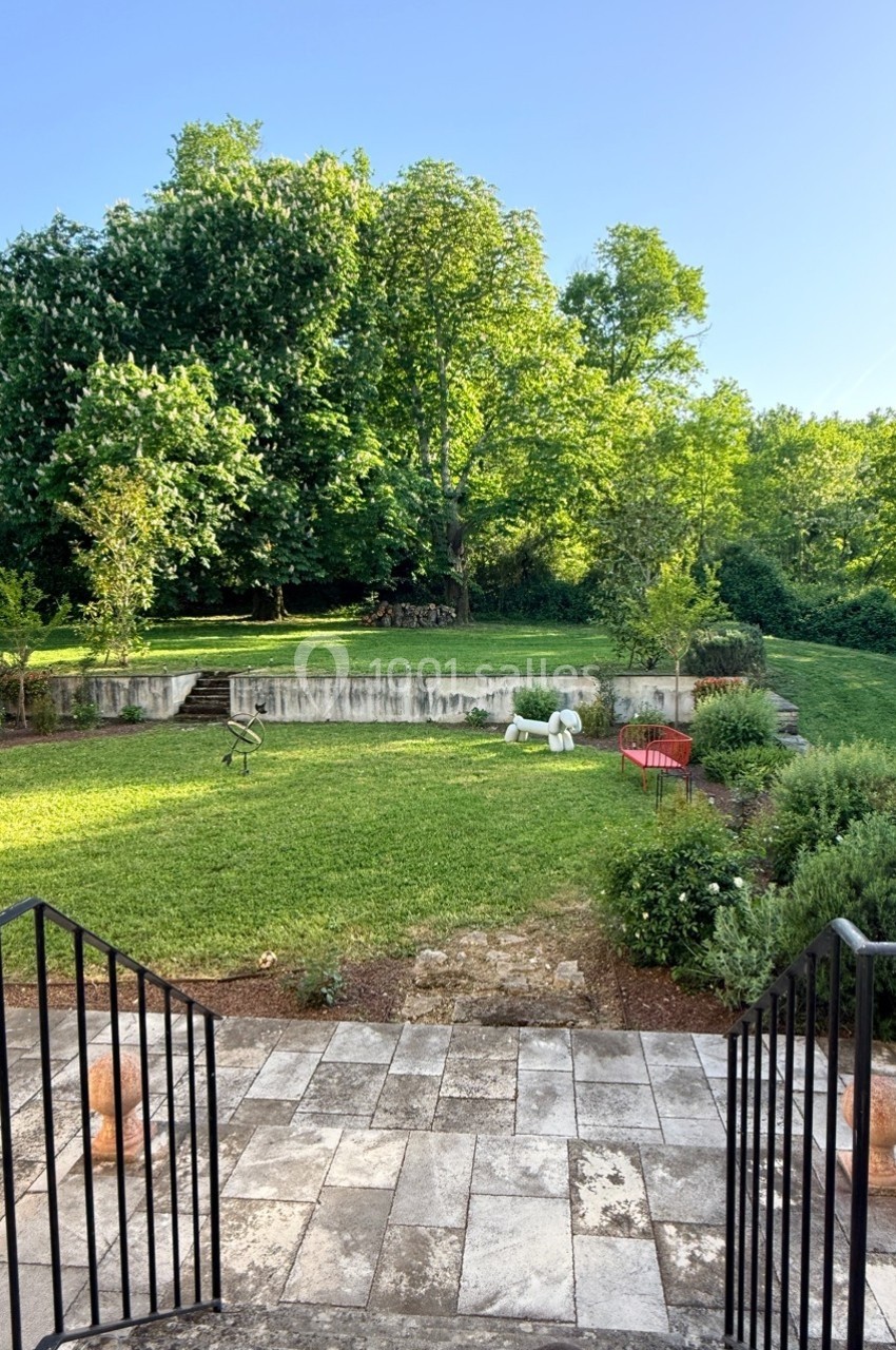 Vue d'un jardin verdoyant avec pelouse, arbres, buissons et une terrasse en pierre au premier plan.