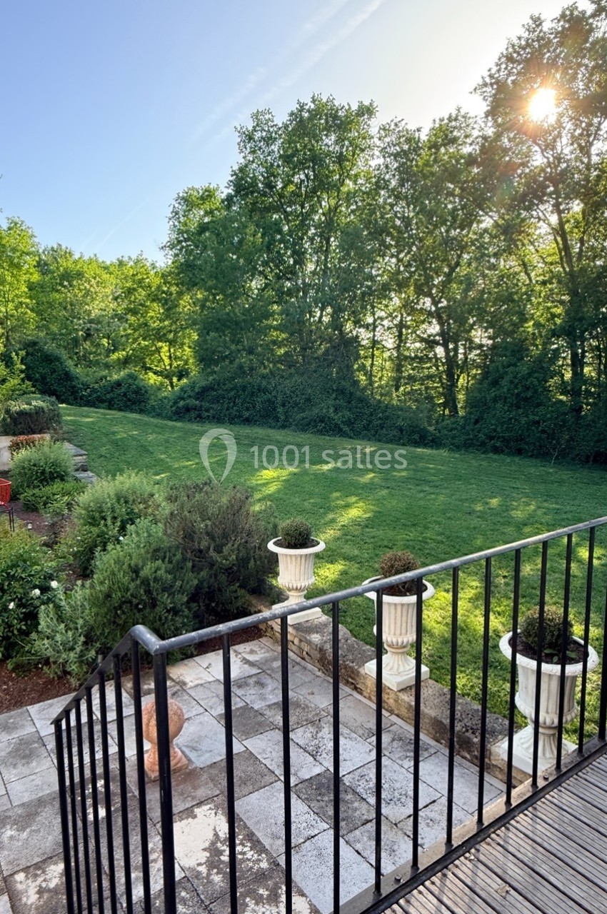 Vue d'un jardin avec pelouse, arbres en arrière-plan, terrasse carrelée et soleil traversant les feuillages.
