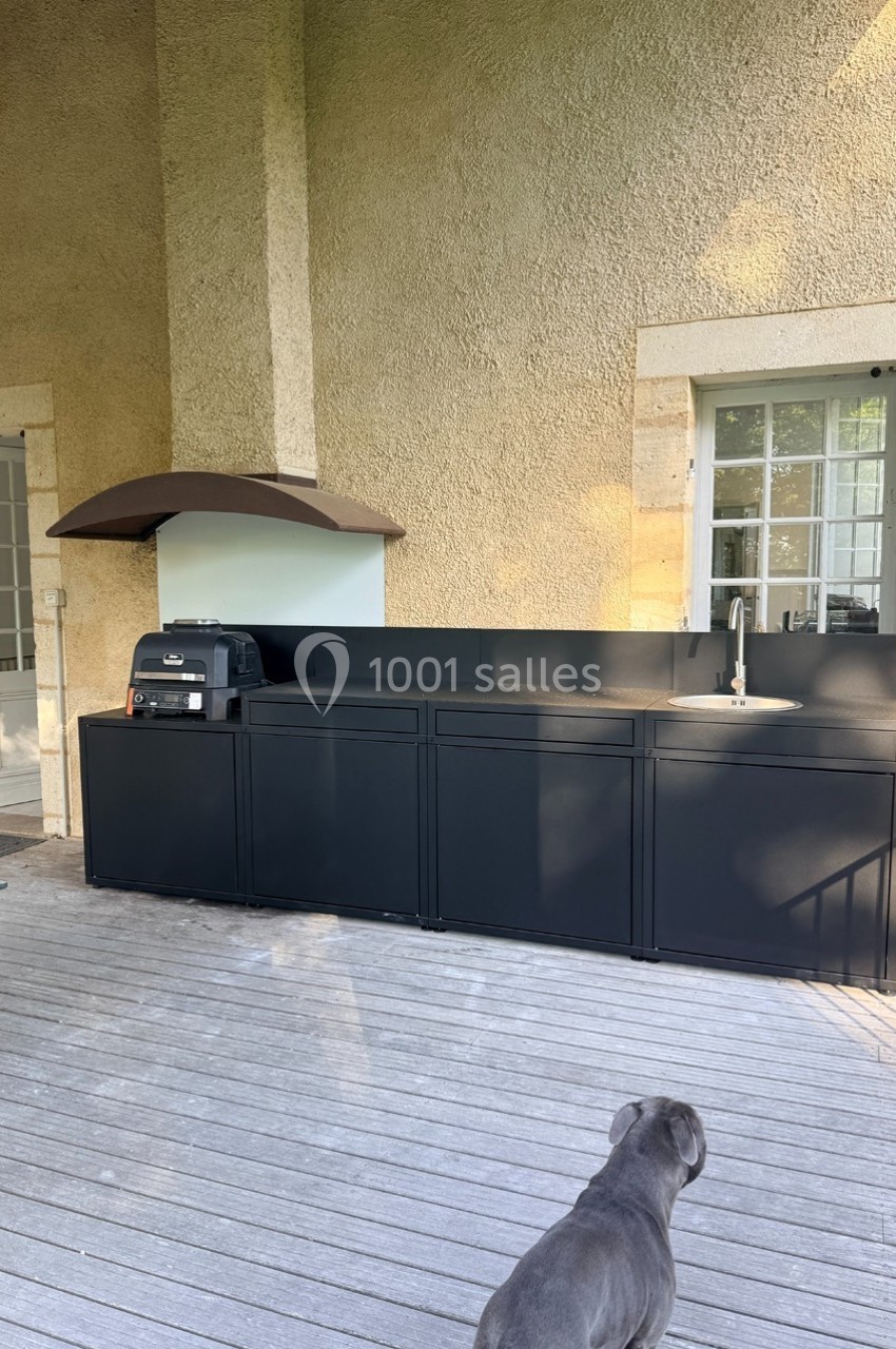 Cuisine extérieure moderne avec plan de travail noir, évier, barbecue et hotte, située sur une terrasse en bois.