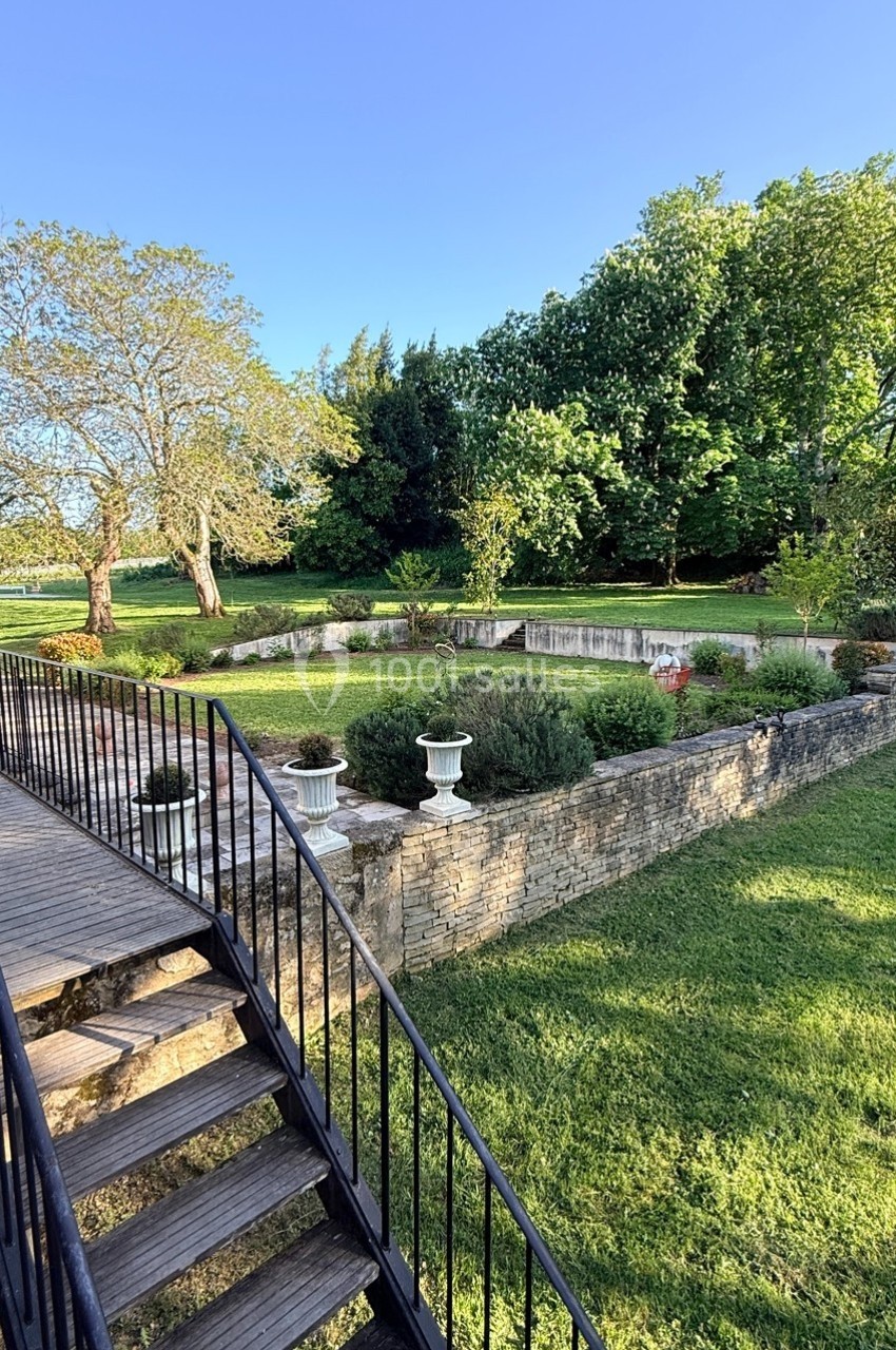 Jardin avec pelouse, arbres, parterre de fleurs surélevé en pierre et escalier en métal menant à une terrasse.