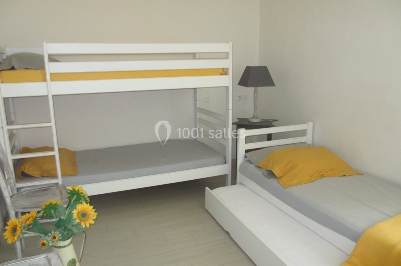 Chambre avec lits superposés blancs, lit simple avec tiroir, linge de lit jaune et gris, lampe et vase de tournesols.